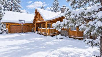 Une cabane en rondins avec un toit enneigé, une terrasse en bois et une cheminée en pierre.