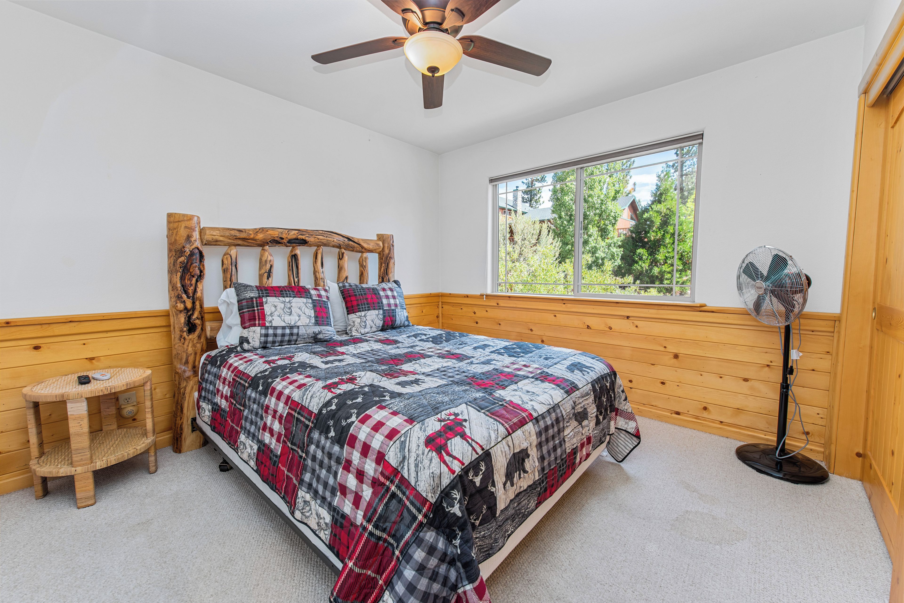 Une chambre avec une tête de lit en bois, une couvre-lit matelassée, un ventilateur sur pied, un petit tabouret en bois et une fenêtre donnant sur des arbres.