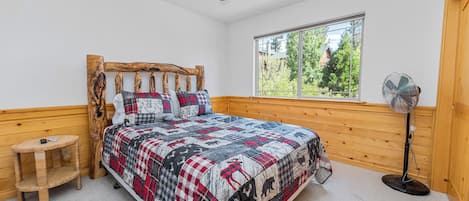 A bedroom with a wooden headboard, a quilted bedspread, a standing fan, a small wooden stool, and a window with a view of trees.