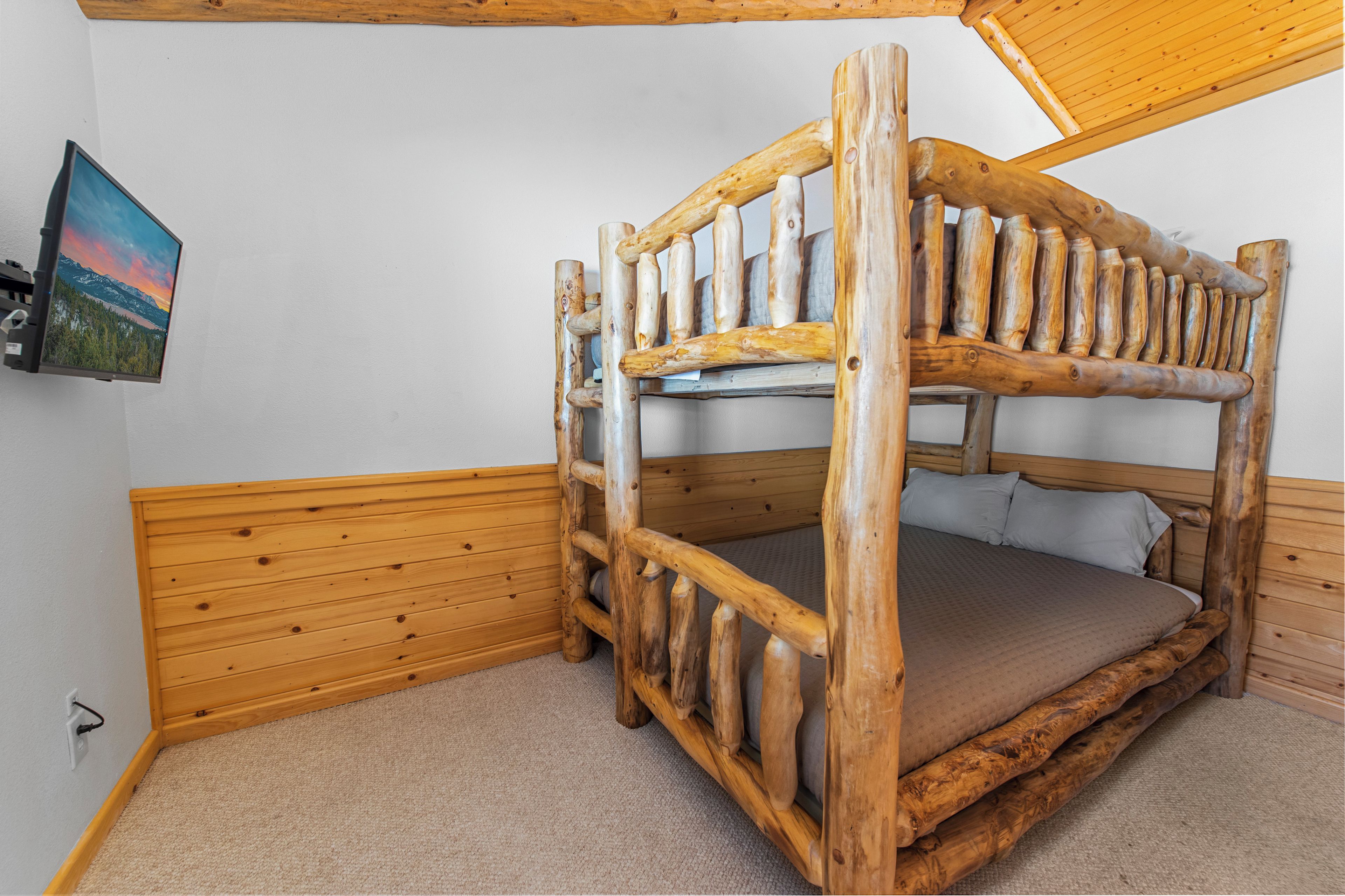 Uma cama beliche com estrutura de madeira, um colchão, uma televisão presa na parede e uma cabeceira de madeira.