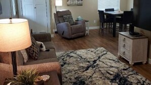 Una sala de estar con sofá, sillón, mesa de centro y mueble para televisión. Hay una zona de comedor con mesa y sillas.