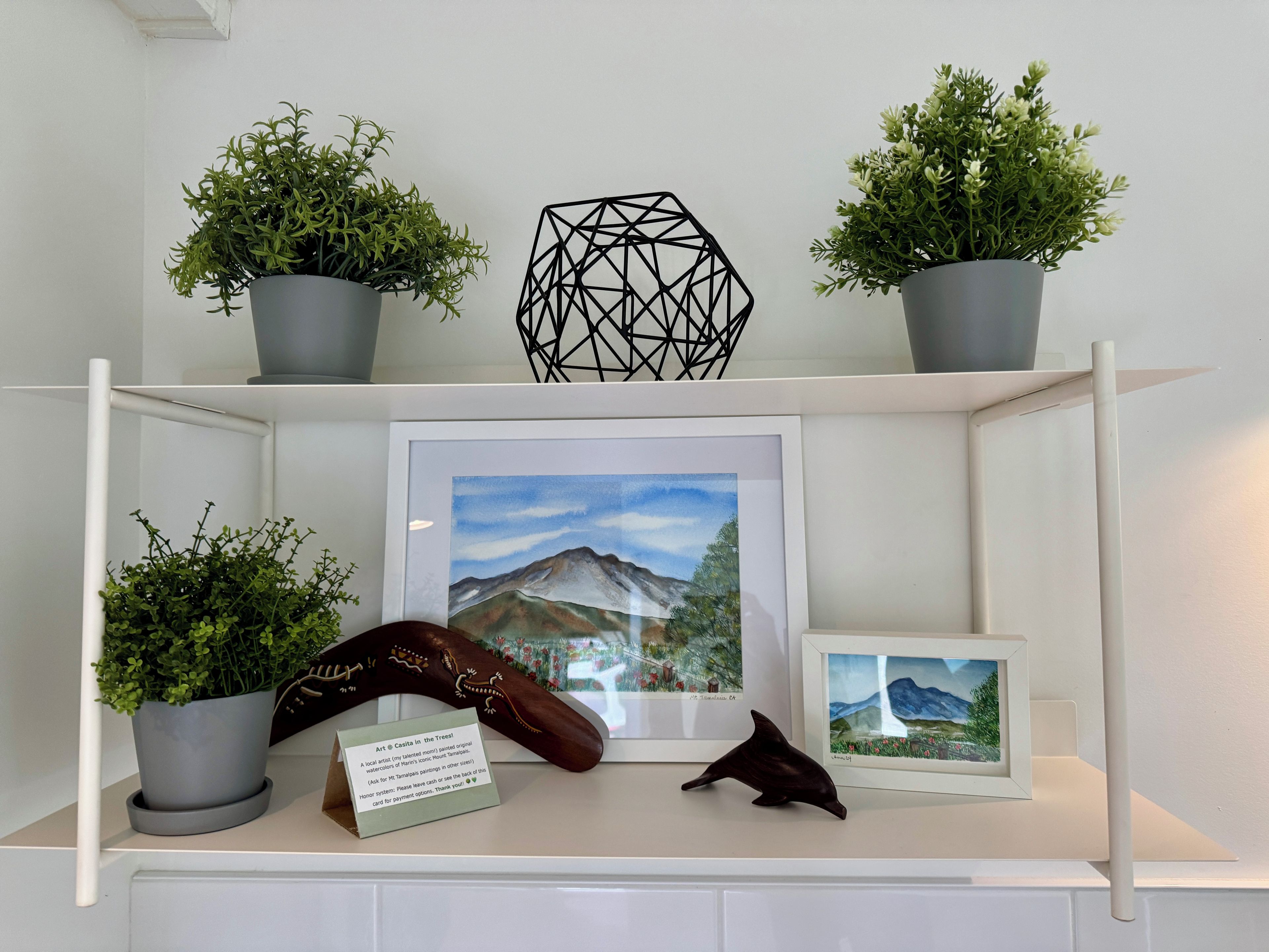 A shelf with potted plants, a framed landscape painting, and a small dolphin figurine.