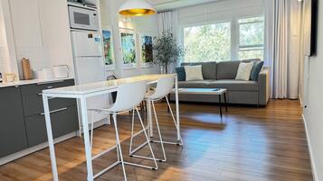 Ett modernt kök med vita skåp, en matplats med vita stolar och en grå soffa i ett vardagsrum med stora fönster.