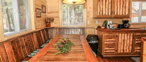 A wooden cabin interior with a long dining table, wooden cabinets, and a window with blinds.