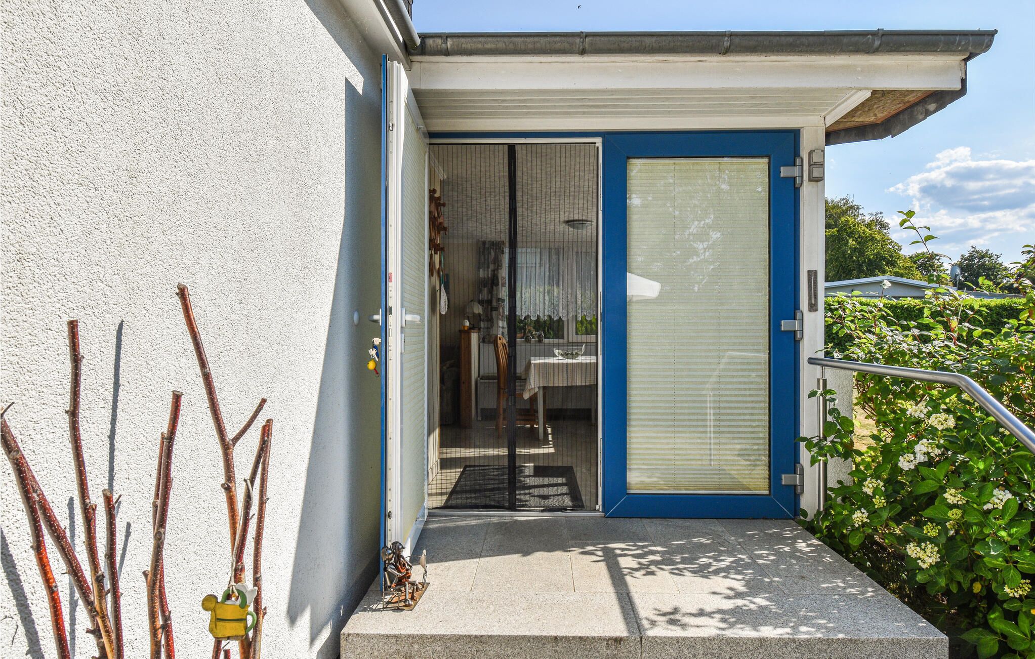 A building entrance with a blue door, a covered outdoor area, and a small garden with plants and a metal railing.
