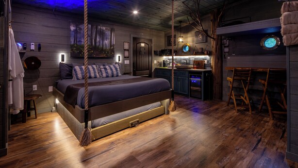 Une chambre avec un lit, un ventilateur de plafond, un plancher en bois, un coin cuisine et une salle de bain.