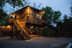 A wooden treehouse with a staircase and a slide, surrounded by trees. - Squirrels Gone Wild Treehouse | 20-Ft Slide, Swings + Rope Climbs (Sevierville)