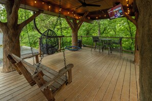 A wooden deck with a swing, a hanging chair, and a table with chairs. - Squirrels Gone Wild Treehouse | 20-Ft Slide, Swings + Rope Climbs (Sevierville)