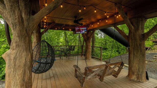 A wooden deck with a swing, hanging chairs, and a ceiling fan. - Family Adventure Treehouse | 20-ft Slide + Secret Beds | Sleeps 6 | Smoky Mtns (Sevierville)