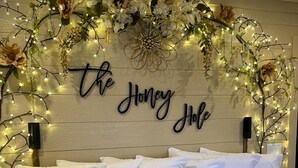 Une chambre avec un lit, du linge de lit blanc, des coussins décoratifs, une enseigne fixée au mur sur laquelle on peut lire « The Honey Hole », et des guirlandes lumineuses.