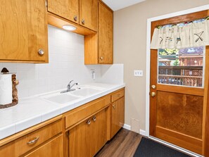 A kitchen with wooden cabinets, a white countertop, a sink, and a door with a window. - Knotty Hideaway (Big Bear City)