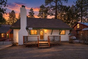A two-story building with a chimney, a porch with a railing, and a garage. - Knotty Hideaway (Big Bear City)