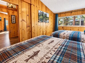 A room with wooden panel walls, a bed with a patterned quilt, a window with a view of trees, and a door leading to another room. - Knotty Hideaway (Big Bear City)