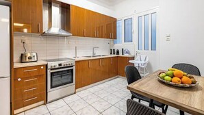 A compact kitchen with wooden cabinets, a stove, and a toaster. There's a dining table with a fruit basket. - Periklis luxury apartment 3 (Athina)