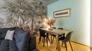 A dining area with a wooden table and chairs, a dark sofa, and a wall mural of palm trees. - Periklis luxury apartment 3 (Athina)