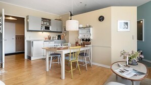 Een moderne keuken met witte kasten, een eettafel met stoelen, een hanglamp en een klok aan de muur.