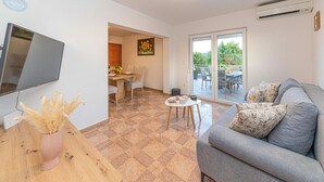 A living room with a flat-screen TV, a sofa, a dining table with chairs, and a balcony with outdoor furniture.
