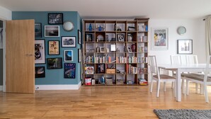 Una habitación con piso de madera, una mesa de comedor blanca con sillas, una estantería llena de libros y objetos decorativos, y una pared azul con cuadros enmarcados.