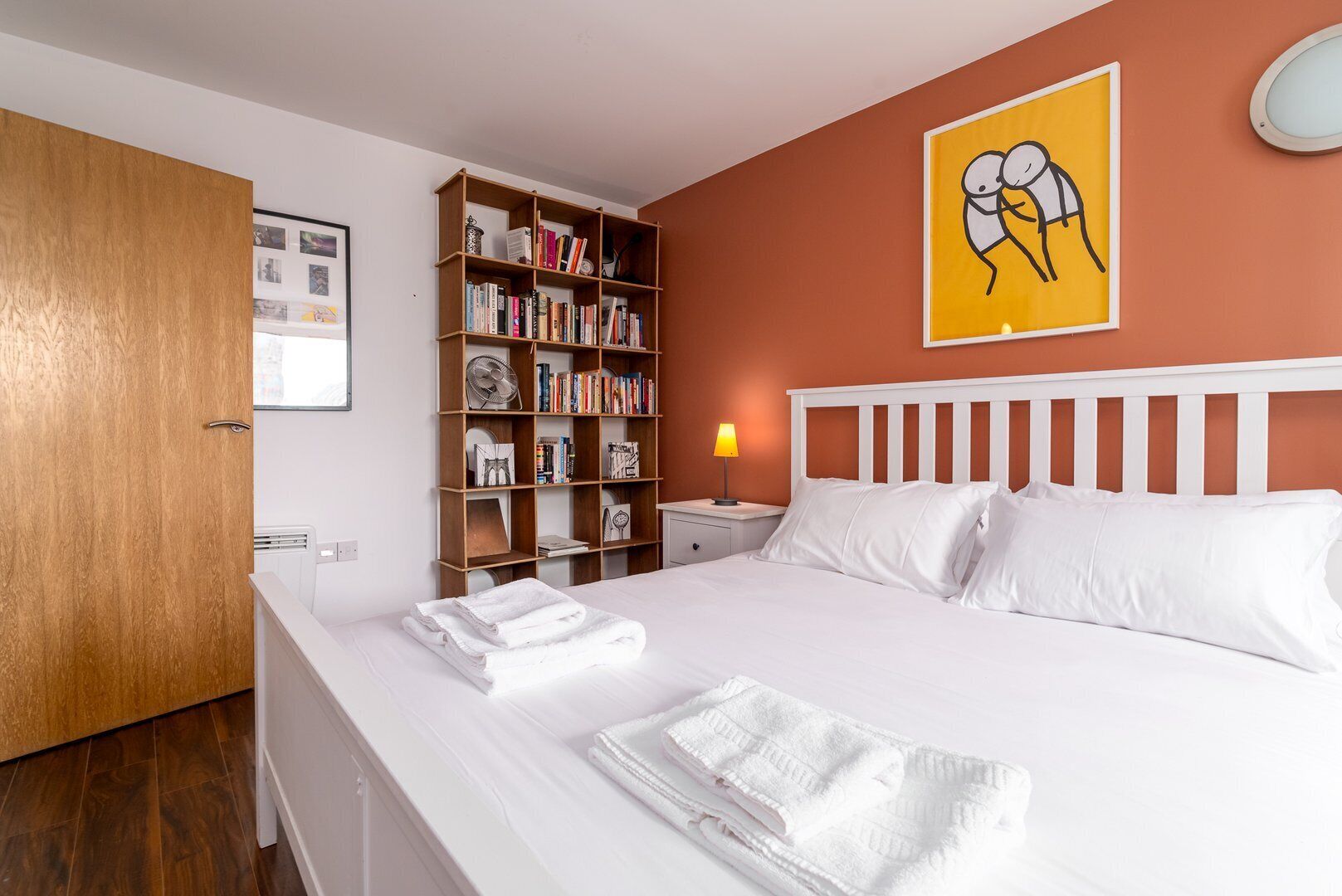 A neatly made bed with white linens, a wooden bookshelf filled with books, a bedside lamp, and a framed picture on the wall.