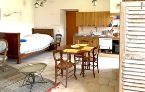 Un dormitorio con una cama, una mesita de noche y una silla. Hay una zona de comedor con una mesa, sillas y un ventanal. Se ve la cocina, con armarios y un lavabo.