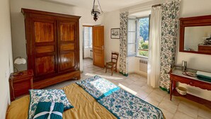 Ein Schlafzimmer mit einem Holzschrank, einem Bett mit floralem Bettbezug, einem Sessel, einem Fenster mit Vorhängen und einem Badezimmer mit Spiegel.