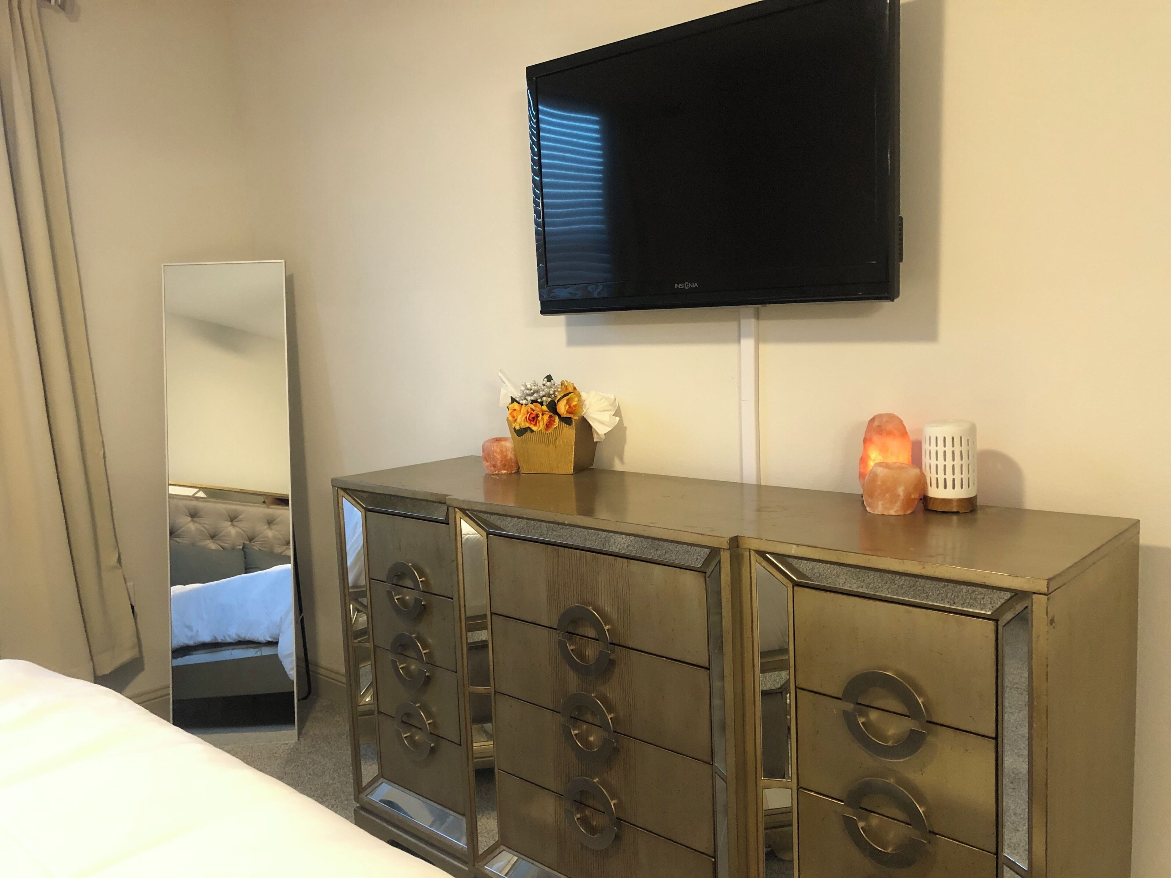 Une chambre d’hôtel dotée d’un grand miroir, d’un tél éviseur fixé au mur, d’une commode avec des tiroirs et d’un lit avec une tête de lit.