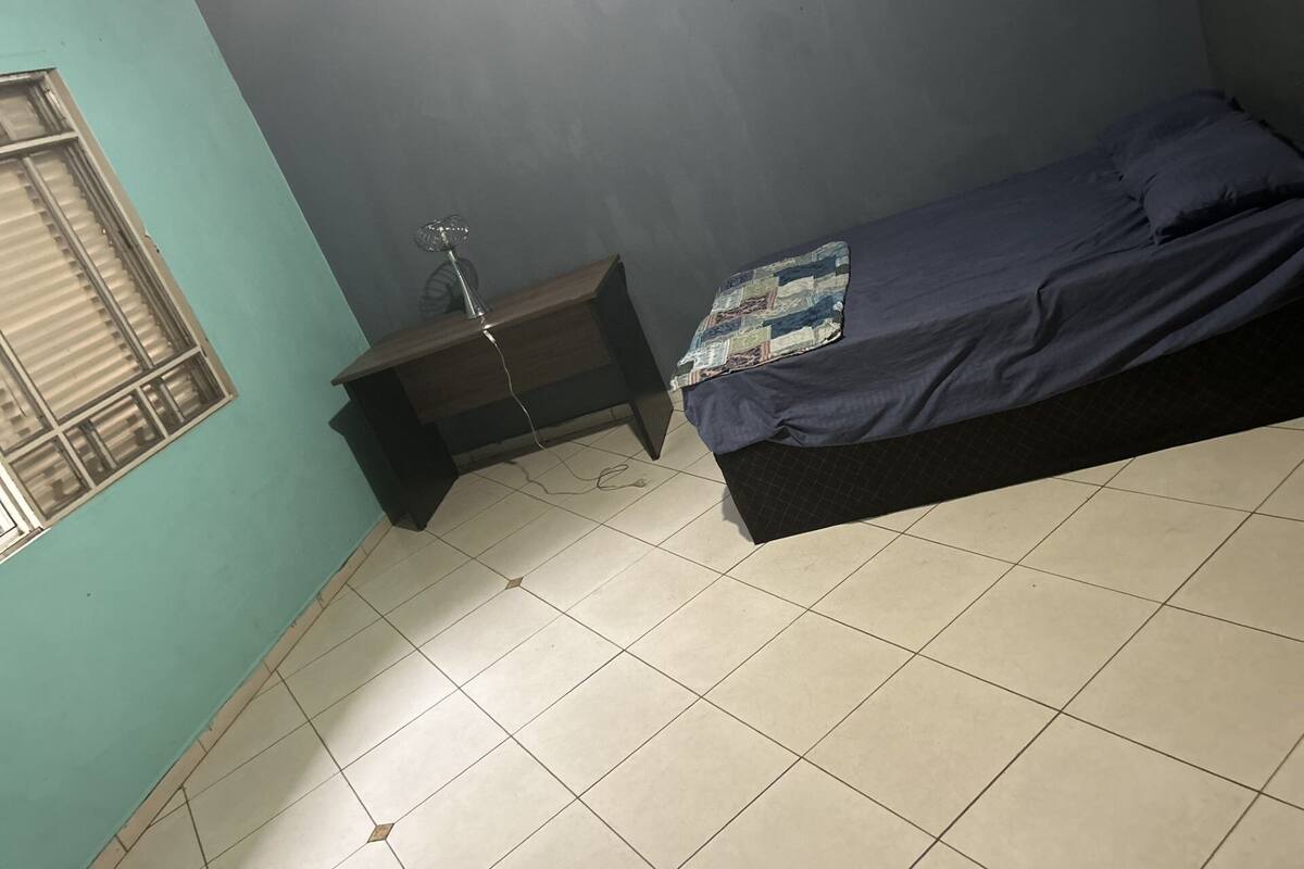 A small, dimly lit room with a single bed, a nightstand, and a window with blinds.