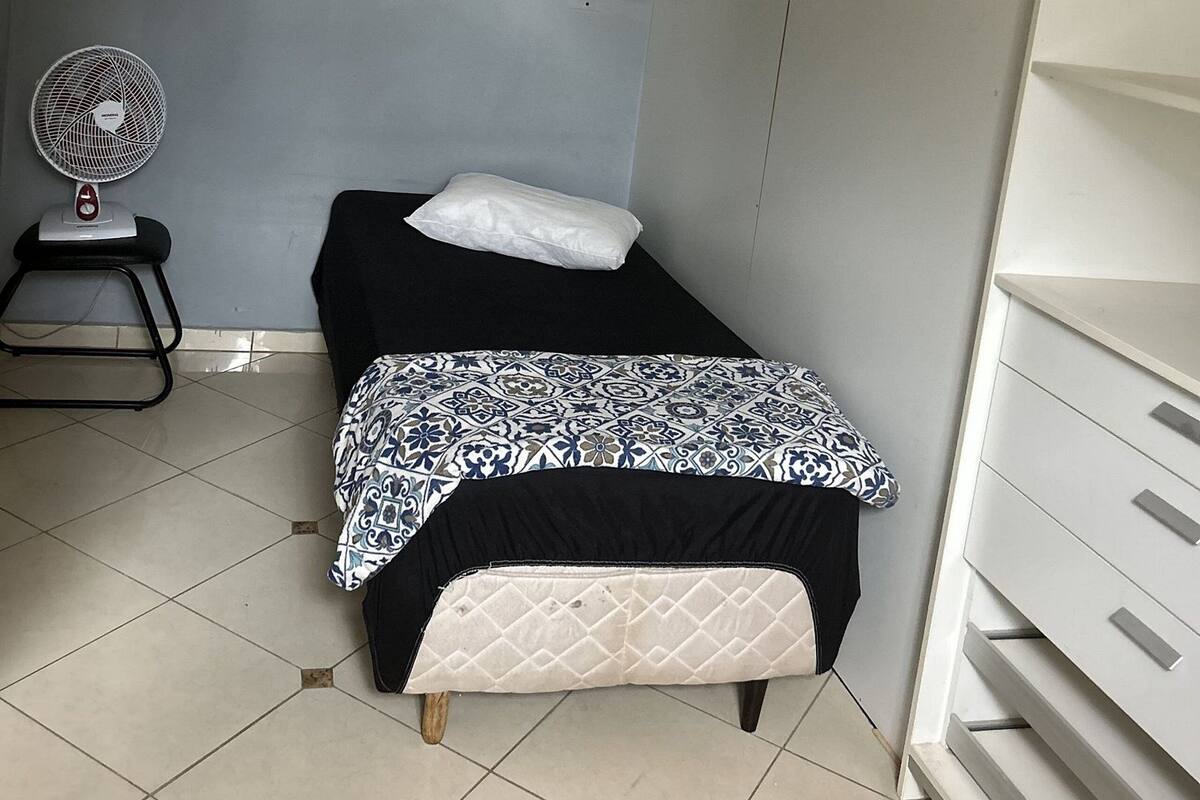 A small, neatly arranged room with a bed, a pillow, a bedside fan, and a chair.