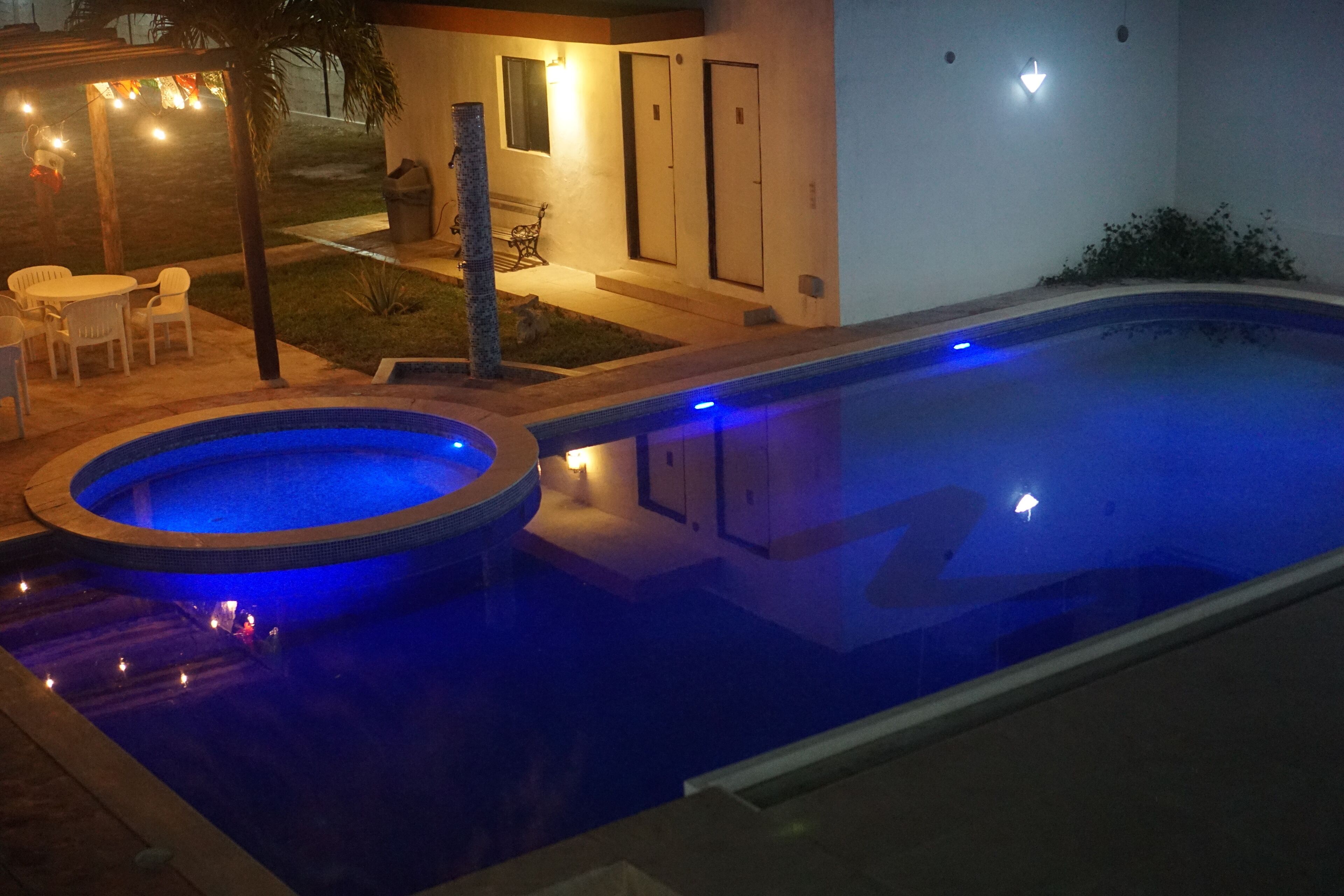 Una piscina con un jacuzzi, rodeada por un patio con sillas y mesas, y un pequeño jardín.