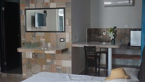 Ett hotellrum med en säng, ett badrum med ett handfat i glas, en spegel och en liten köksdel med en stol och en vas med blommor.