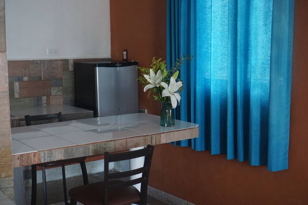 A dining area with a table, chairs, a vase with flowers, a refrigerator, and blue curtains.