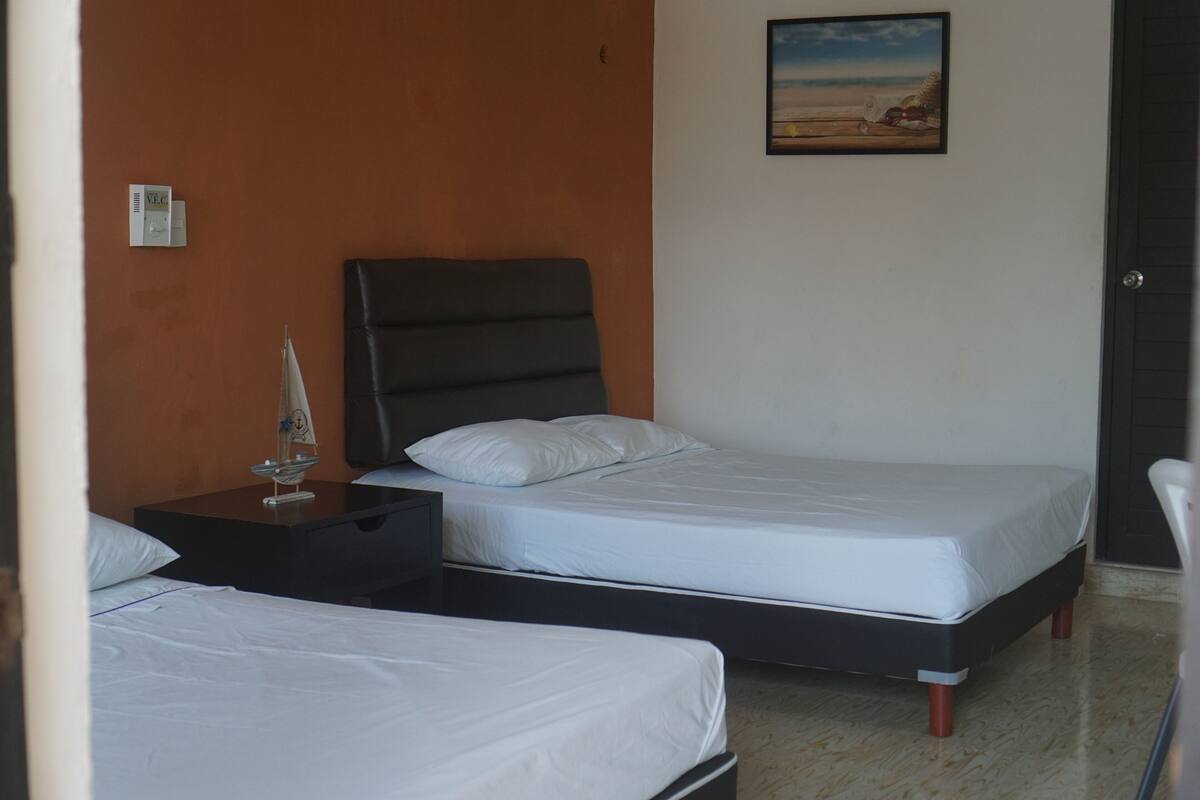 A hotel room with two single beds, a nightstand with a lamp, a painting on the wall, and a door.