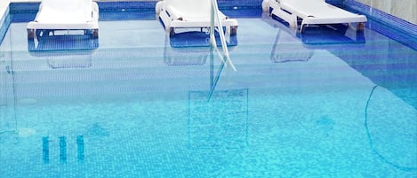Una piscina con tres sillas reclinables blancas y un suelo de baldosas azules claras.