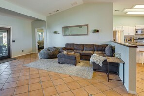 Ein Wohnzimmer mit einem Ecksofa, einem Sitzsack, einem kleinen Hocker und einem Couchtisch. Im Hintergrund ist eine Küchenzeile mit Schränken und Geräten zu sehen.