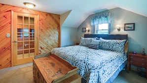 A bedroom with a wooden door, a bed with patterned bedding, a bedside table with a lamp, and a nightstand with a phone. - Spacious Lowville Retreat on 4 Private Acres! (Lowville)