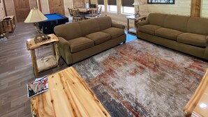 Ein geräumiges Wohnzimmer mit Holzwänden und -decke, einem Billardtisch, zwei Sofas und einem Deckenventilator.