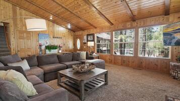Interior acogedor de una cabaña con un techo de madera grande, un sofá modular, una mesa de centro con objetos decorativos y ventanas amplias que ofrecen vistas al exterior.