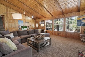 Un intérieur de chalet chaleureux, avec un grand plafond en bois, un canapé d’angle, une table basse agrémentée d’objets décoratifs et de grandes fenêtres offrant une vue sur l’extérieur.