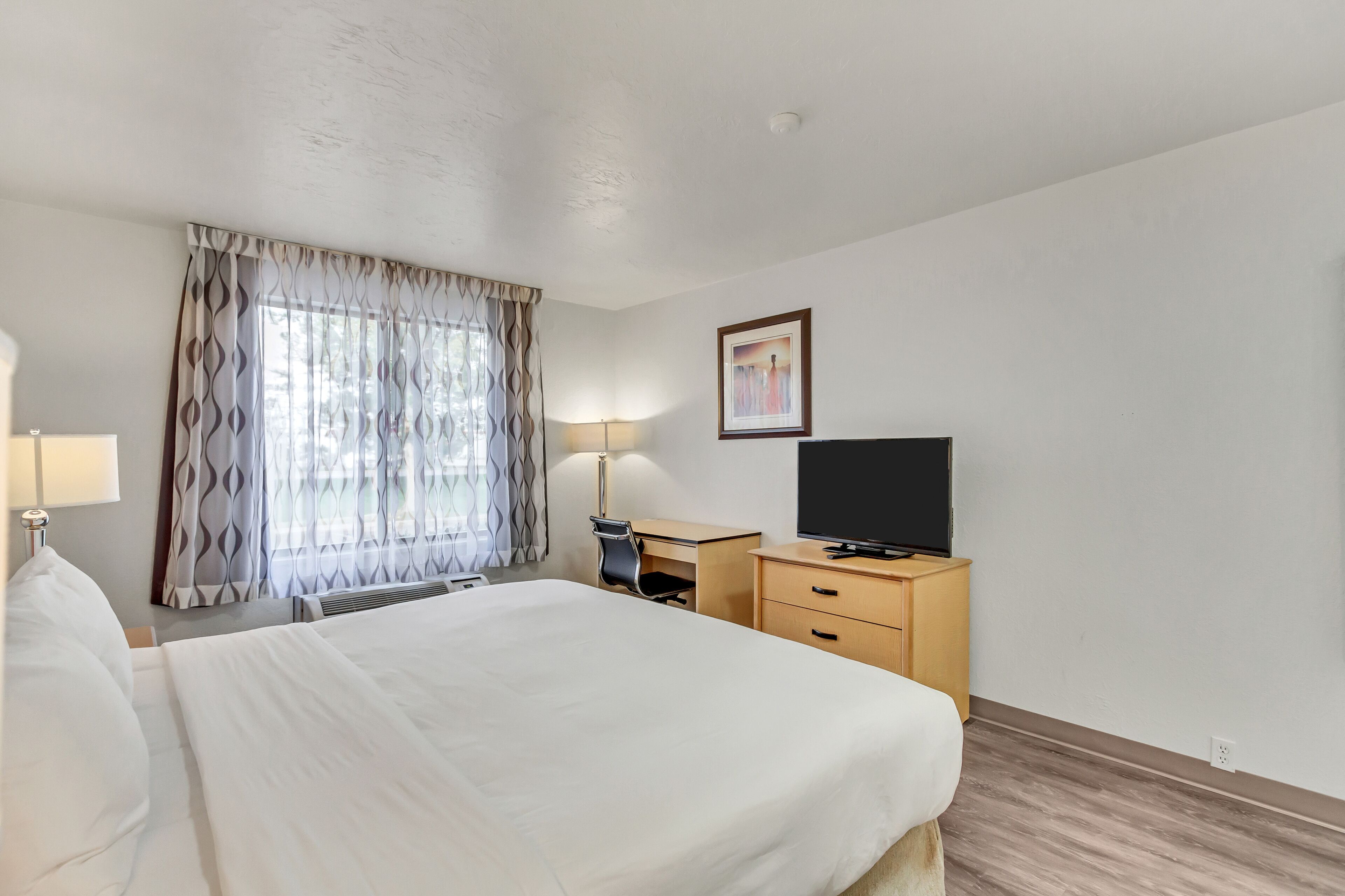 Quarto de hotel com cama, escrivaninha, cadeira, televisão e janela com cortinas.