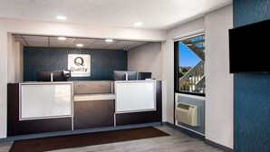 Ein Empfangsschalter mit einem 'Quality by Choice Hotels'-Schild, einem Flachbildfernseher und Blick auf eine Treppe sowie Grünflächen im Außenbereich.