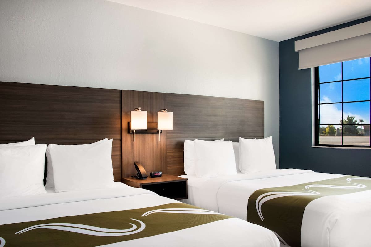 Une chambre d’hôtel avec deux lits, une table de chevet avec une lampe et une fenêtre donnant sur le ciel et les arbres.