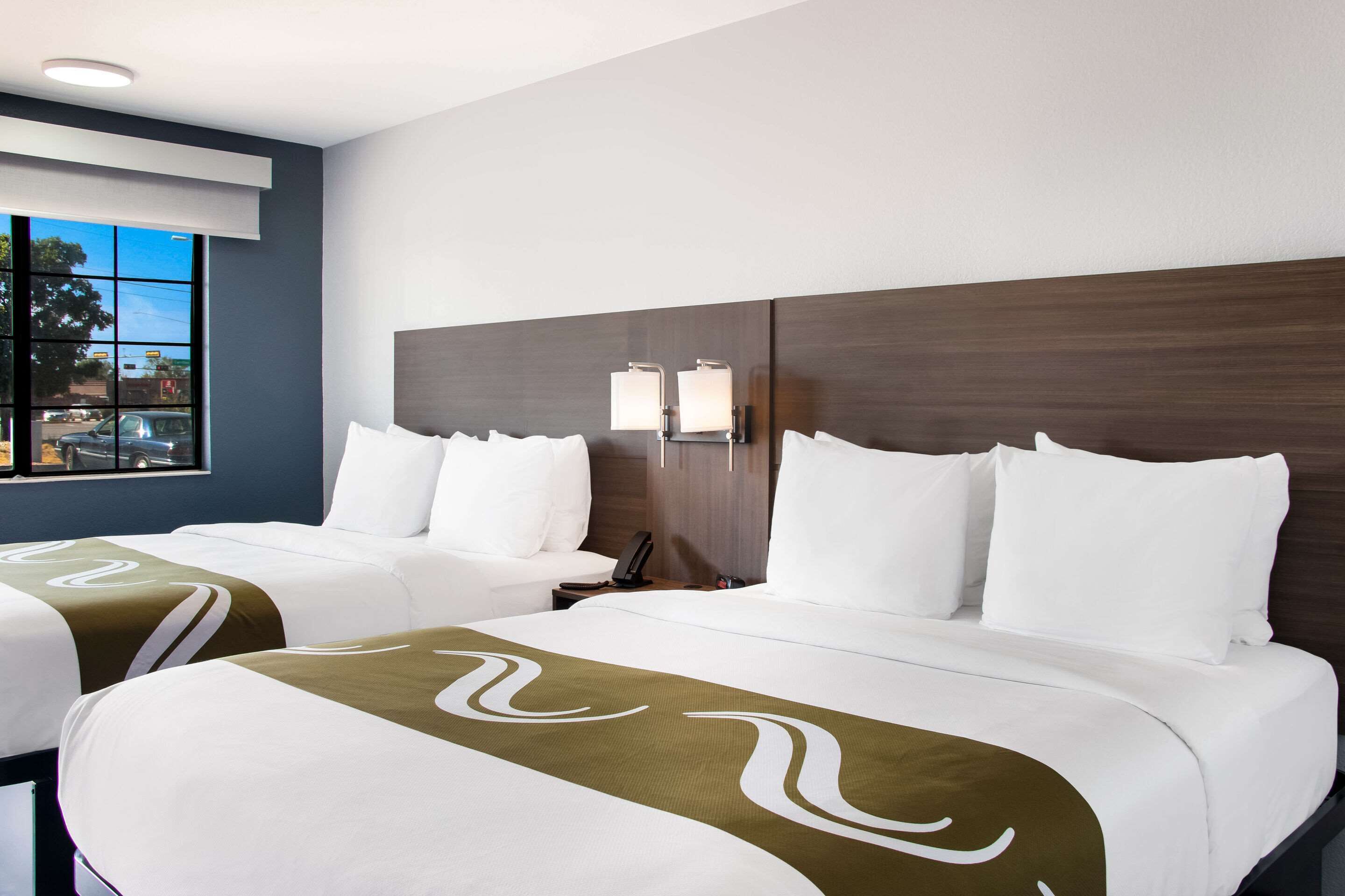 A hotel room with two beds, a large window, a nightstand, and a wall-mounted lamp.