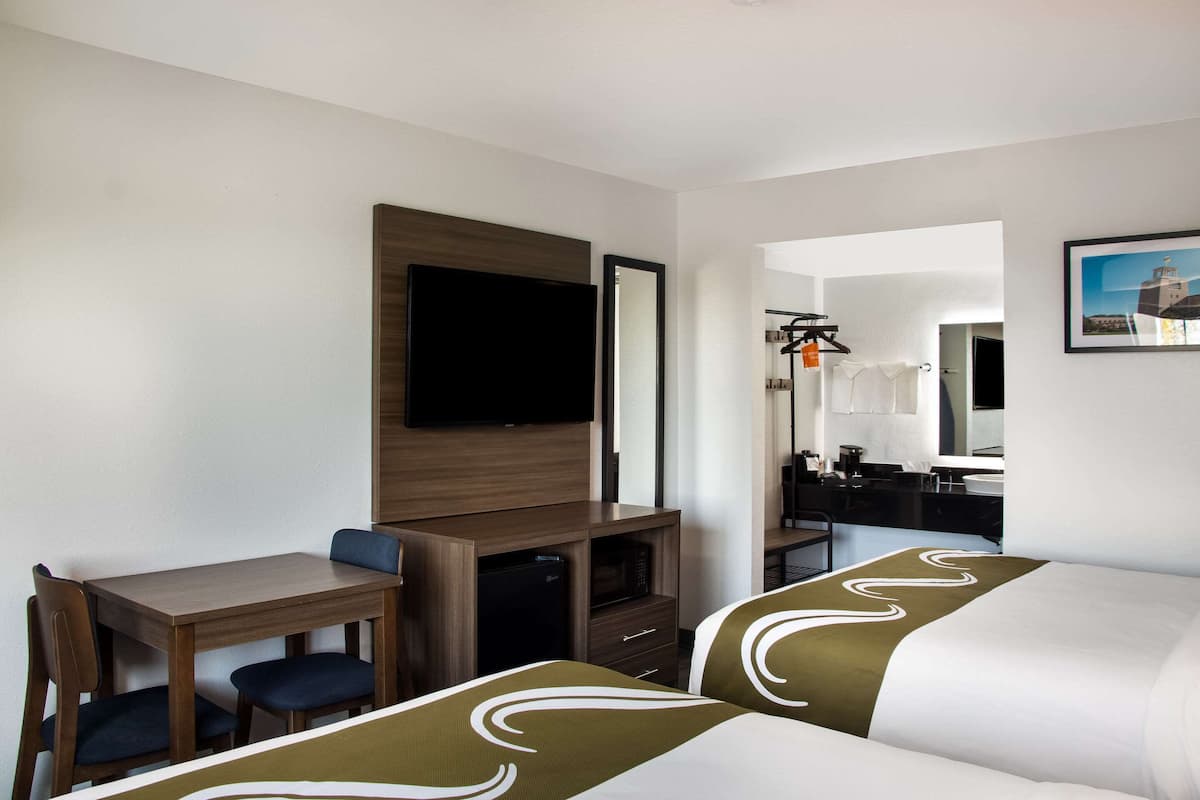 Une chambre d’hôtel avec un lit, un bureau, une chaise, une télévision, un miroir et une salle de bain visible à travers une porte ouverte.
