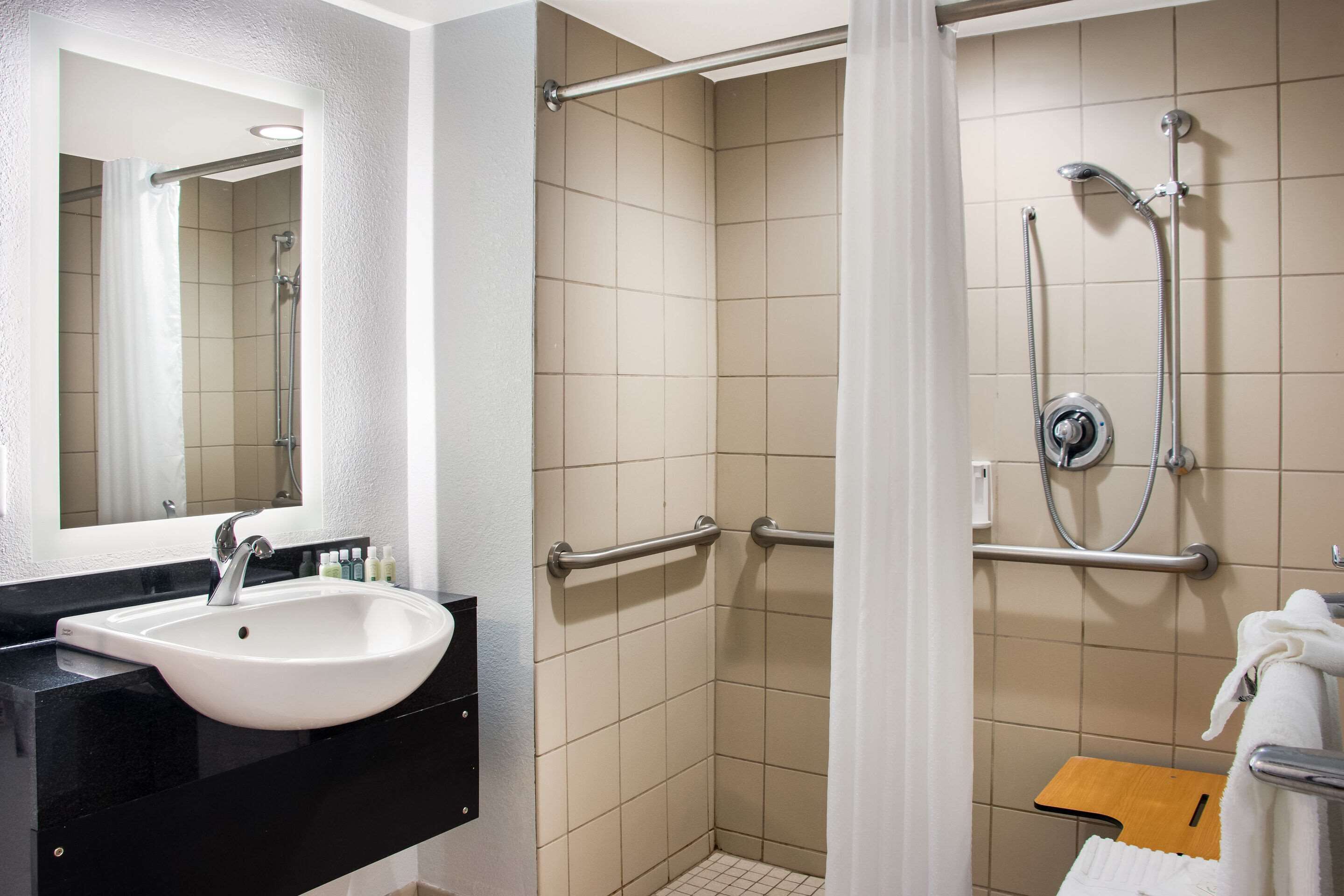 Un bagno con un lavandino bianco, uno specchio, una doccia dotata di maniglie di sostegno e un piccolo sgabello di legno.