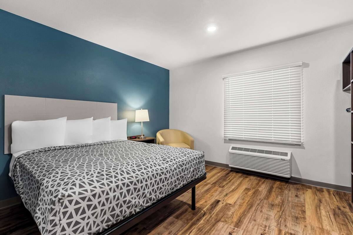 Una habitación de hotel con una cama, una silla, una mesita de noche, una lámpara y una ventana con persianas.