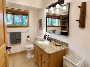Une salle de bain avec des toilettes blanches, un lavabo sur un plan de travail en granit, un miroir et une fenêtre avec un porte-serviettes.