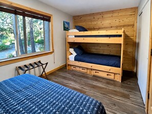 A room with a bunk bed, a single bed, a window with a view of trees, and a wooden wall.