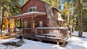 A wooden cabin with a porch and deck, surrounded by trees and snow. - Adorable Cabin 5-min walk to beach! Dog friendly! (Tahoe Vista)