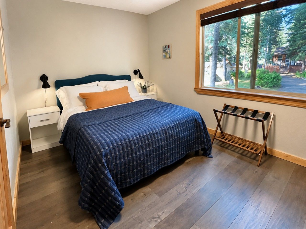 A bedroom with a bed, a nightstand, a lamp, a window with a view of trees, and a wooden rack.