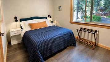 A bedroom with a bed, a nightstand, a lamp, a window with a view of trees, and a wooden rack.